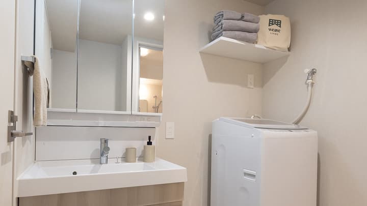 Modern washroom with a vanity sink, large mirrored cabinet, washing machine, and a shelf with towels and a storage bag.