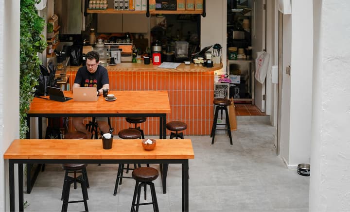 A man working on a laptop at a long wooden table in a brightly lit coffee shop or common area with an orange tiled counter and stools in the background.