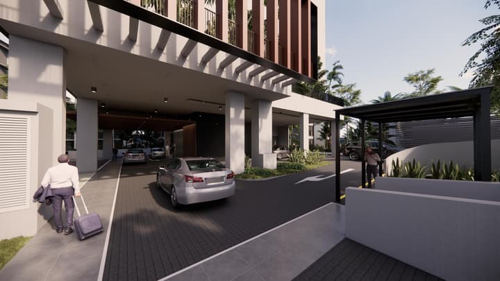 The covered entrance and drop-off area of a modern building, showing a security gate, cars, and a pedestrian pulling luggage, framed by concrete columns and landscaping.