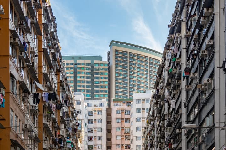 View looking up between two tall, densely packed apartment buildings with numerous exterior air conditioning units and hanging laundry, focusing on modern high-rises in the distance against a blue sky.