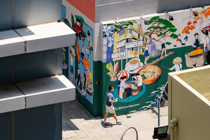 A man walking past a vibrant, large outdoor street art mural depicting scenes of everyday Hong Kong city life and culture on the side of a building.