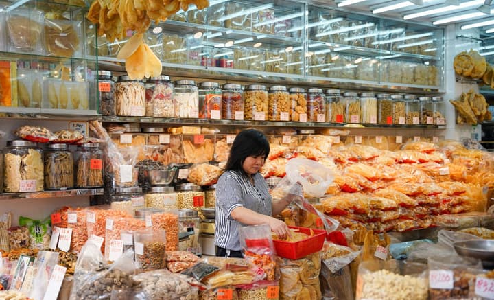 A woman working at a stall in a traditional Hong Kong dried goods market, surrounded by jars and bags of various dried seafood, nuts, and snacks.