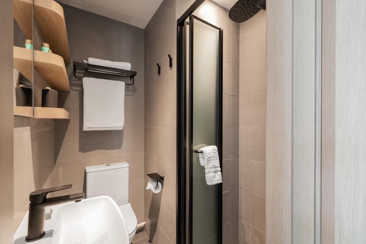 A compact, contemporary bathroom with neutral gray tile walls, a white sink and toilet, a black-framed glass shower door, and floating light wood shelves.