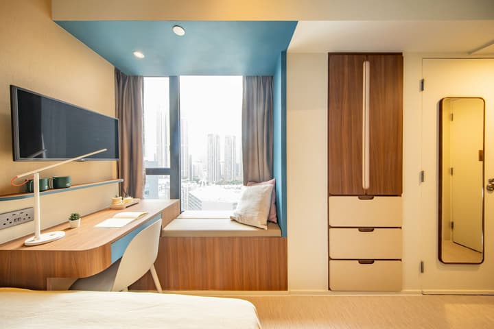 A close-up view of the study area in a small studio, showing a wood-top desk with a light blue panel next to a window seat. The desk has a white lamp and a wall-mounted TV. To the right, there is a tall, narrow storage cabinet with light-colored drawers and a full-length mirror mounted on a nearby door.