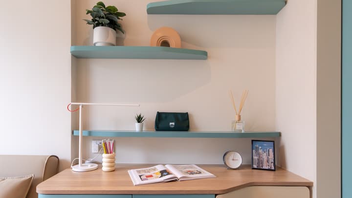 Close-up of a wooden desk and white wall with two floating mint-green shelves, decorated with a lamp, plants, books, and small decorative items.
