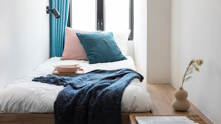 A close-up, minimalist view of a single bed in a tight nook, dressed with a dark blue knitted throw blanket, a pink pillow, and a teal textured pillow, next to a window with bright blue curtains.