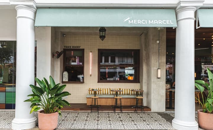 The modern cafe facade of Merci Marcel, featuring white columns, a pale green awning, checkered tile flooring, and outdoor bench seating with small tables.
