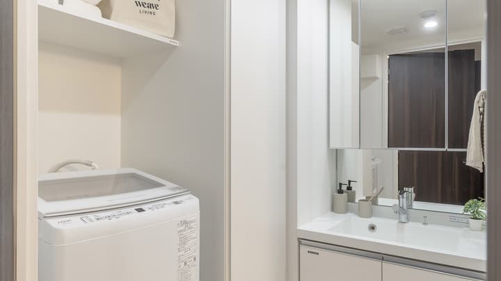 Laundry alcove with an AQUA top-loading washing machine tucked beneath a shelf, next to the modern bathroom vanity with mirrored medicine cabinets.