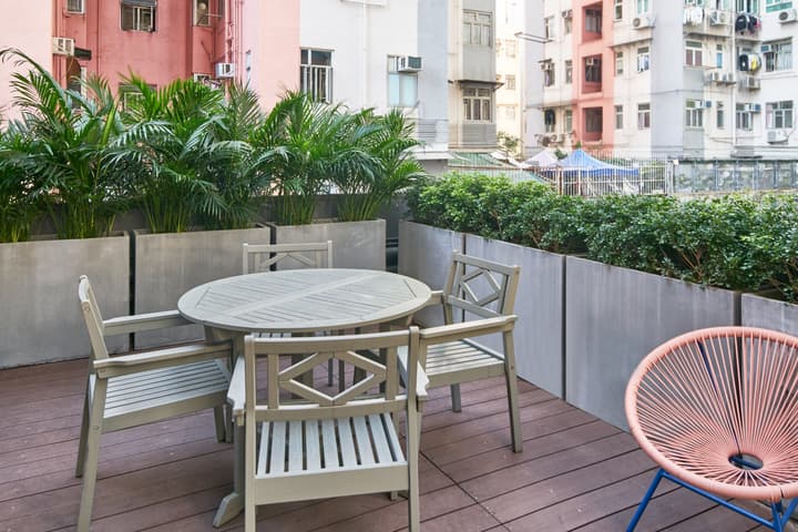 Outdoor rooftop terrace with a wooden-slat deck, featuring a round gray wooden dining set surrounded by planters with dense green foliage against a backdrop of colorful residential buildings.