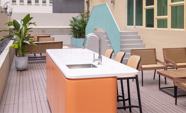 An outdoor patio area with wood decking, featuring a modern cooking island with a sink and an orange base, surrounded by black and rattan-style bar stools and outdoor furniture.