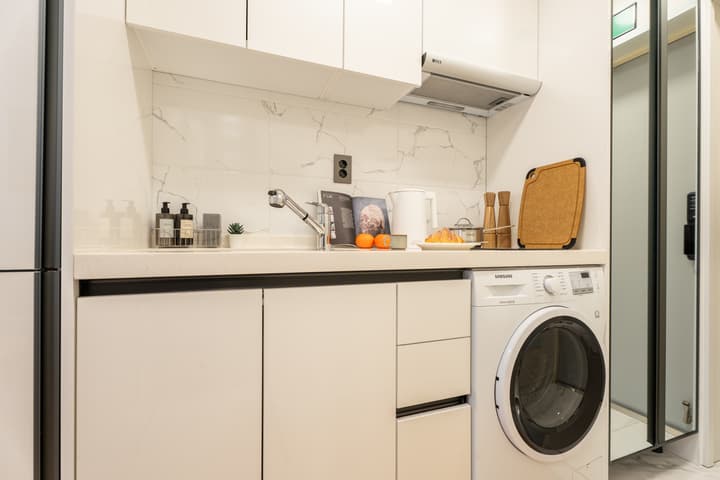 Clean, white kitchen with a marble-effect backsplash, under-cabinet storage, a sink, and a built-in Samsung washer/dryer combo beneath the counter.