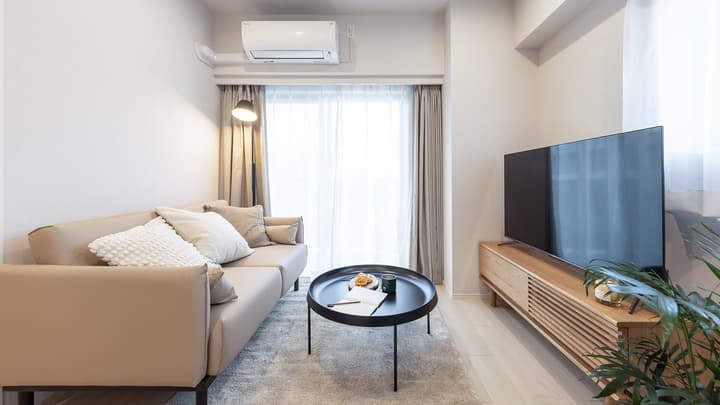 Bright living room with a neutral-toned sofa, black round coffee table, a light wood TV console, a large TV, and floor-to-ceiling sheer curtains over a window or balcony door.