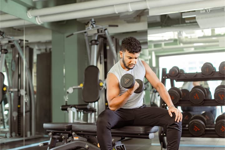 A focused young man sitting on a weight bench and performing bicep curls with a dumbbell in a brightly lit modern fitness center.