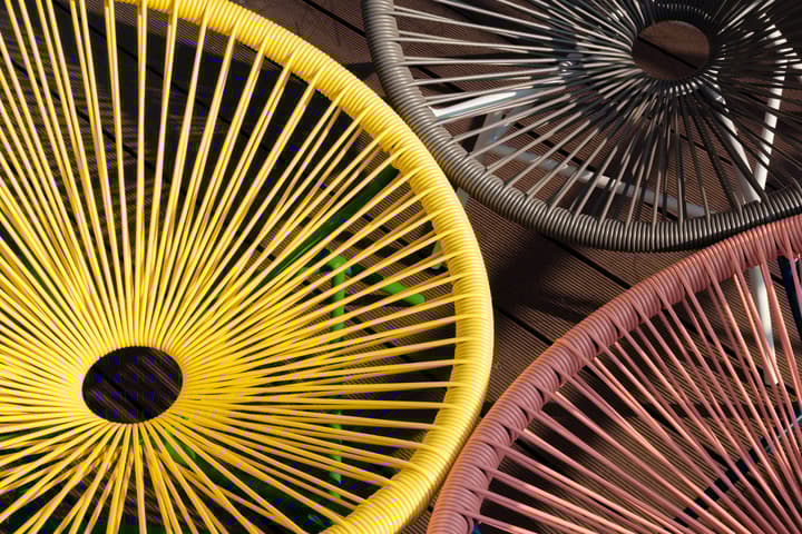 Abstract close-up, overhead view of the woven-spoke seats of three brightly colored Acapulco-style outdoor chairs in yellow, charcoal gray, and dusty pink, set on a dark wood-plank deck.
