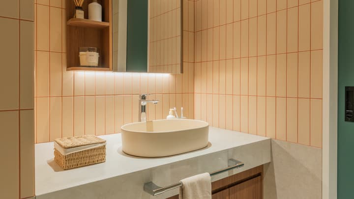 Modern bathroom vanity area with a beige oval vessel sink on a light gray countertop, mirrored medicine cabinet, and walls tiled with vertical pale pink/orange tiles.