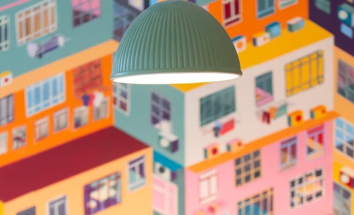 Close-up view of a ribbed, domed green light fixture hanging in the foreground, with an out-of-focus background showing a brightly colored, isometric mural of apartment buildings.