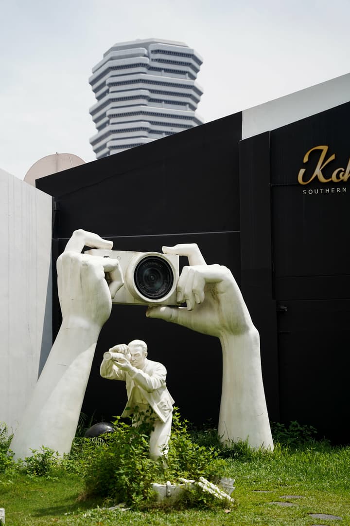 Outdoor art installation featuring large white sculpted hands holding a camera, with a smaller figure below, set against a black building facade and a modern stacked high-rise in the background.
