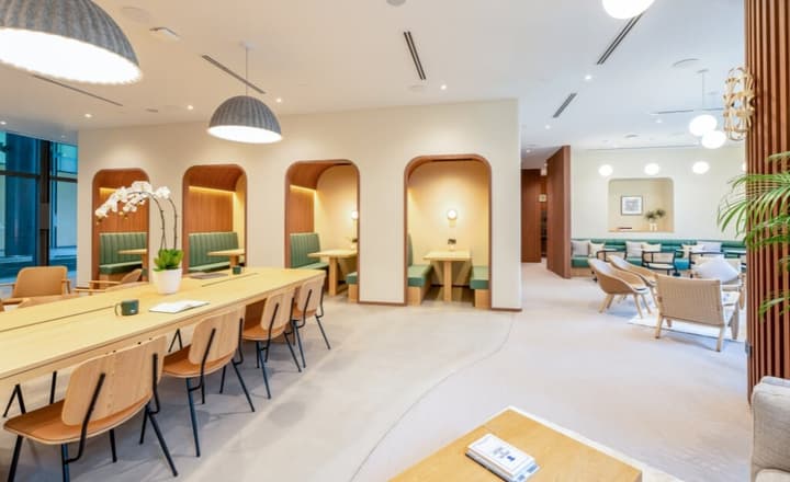 Wide view of the co-working lounge with individual green upholstered booths with wood arches on the left and a large communal wooden table on the right.