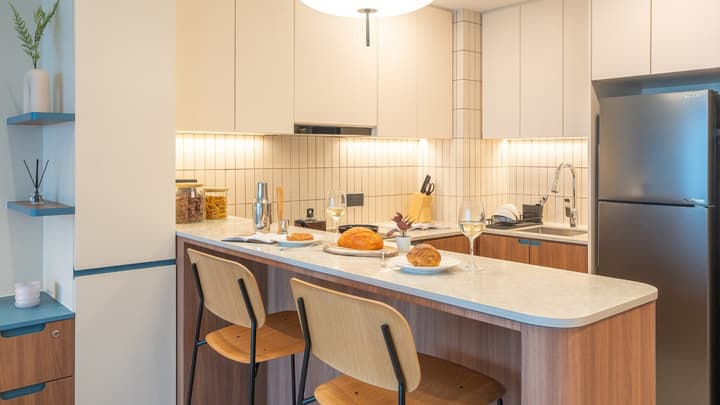 Compact kitchen featuring a breakfast bar with two wooden stools, a light countertop with breakfast items, and white and wood-grain cabinets.