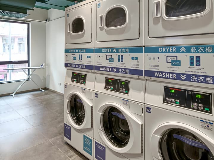 Clean, brightly lit shared laundry room with a stack of commercial washers and dryers, a large window, and an ironing board setup.