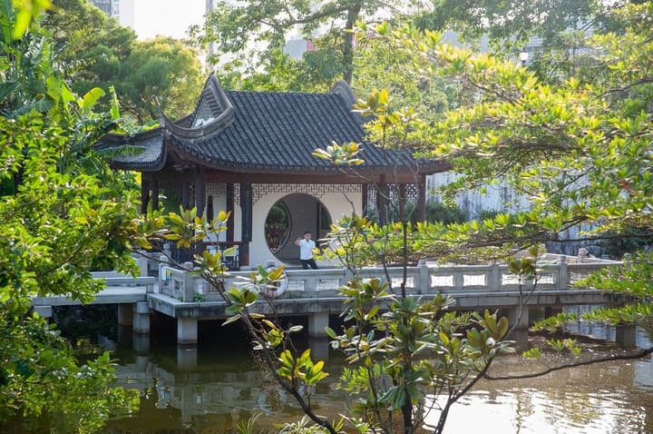 Traditional Chinese garden scene with a dark-roofed pavilion, round moon gate, and stone bridge over a pond, surrounded by lush green trees and foliage. 