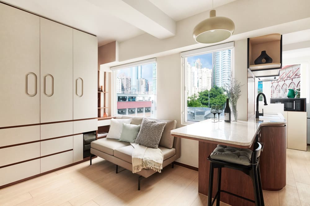 Contemporary, open-plan studio apartment featuring a sofa, sleek built-in floor-to-ceiling storage cabinets, a kitchenette with a breakfast bar, and large windows with a city view.