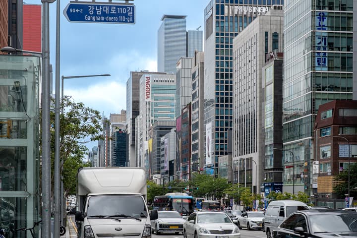 A view of the Gangnam-daero road in Seoul, with a sign for 'Gangnam-daero 110-gil' and a dense skyline of modern, glass high-rise commercial buildings.