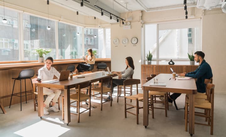 Bright, modern coworking space with large windows overlooking a city view, featuring long communal wooden tables, mid-century style chairs, and several people working on laptops and reading.