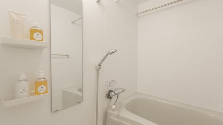 View inside a bright, white Japanese-style bathroom featuring a bathtub, a shower head, and a large mirror with shelves holding toiletries.
