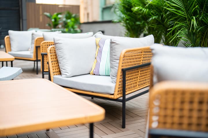 Close-up of comfortable outdoor patio seating featuring chairs with woven rattan-look sides, black metal frames, and thick gray cushions. Tropical plants provide a lush background, and wooden tables are visible in the foreground.