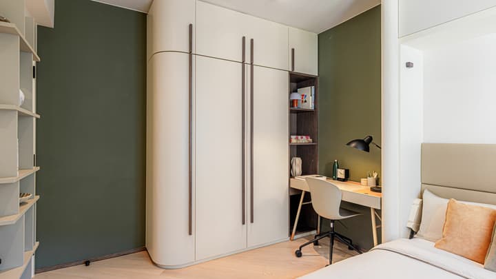 Flexible apartment room set up as a bedroom with a desk, built-in curved wardrobe, and a dark green accent wall behind the desk area.