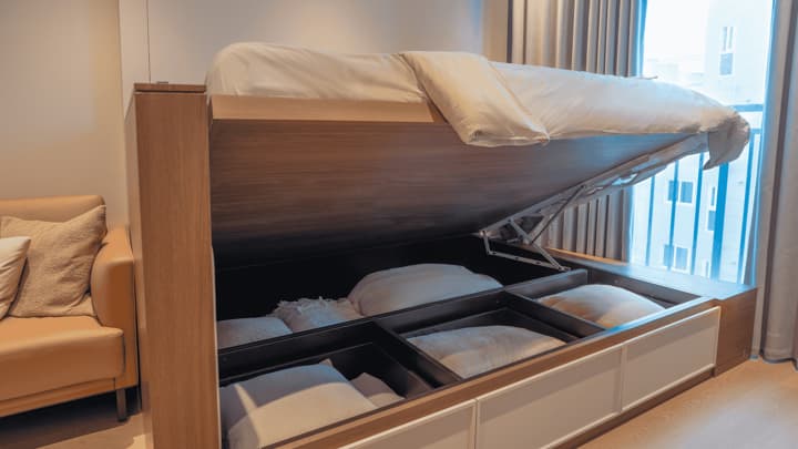 A full-size bed lifted on a hydraulic mechanism to reveal deep, segmented storage compartments filled with extra pillows and linens.