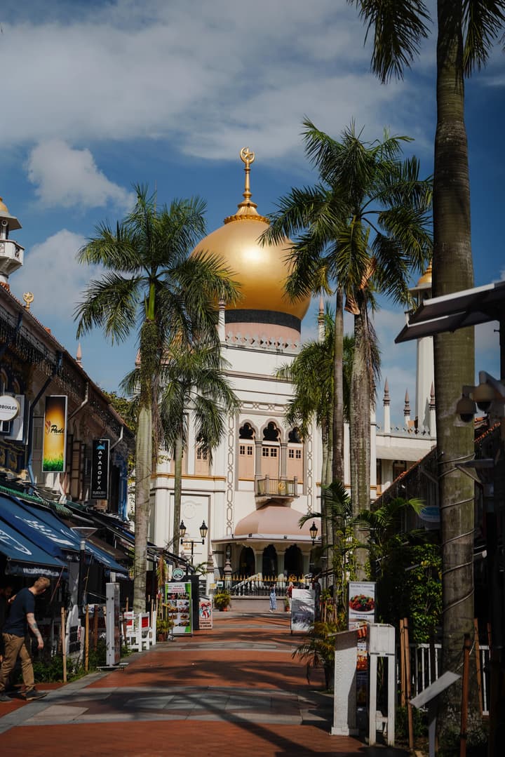 Scenic view down a pedestrian street in Kampong Glam, Singapore, featuring tall palm trees and the majestic Sultan Mosque with its striking gold dome.