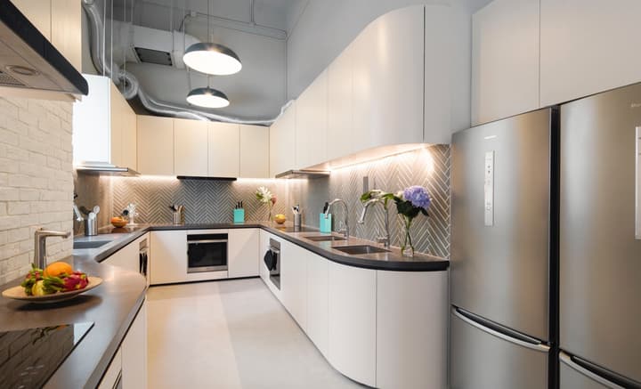 A modern, high-end communal kitchen with a U-shaped counter, white curved cabinets, stainless steel appliances, and a gray herringbone tile backsplash with under-cabinet lighting.