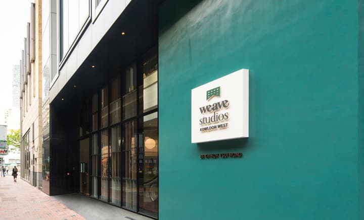 Exterior view of a modern building entrance. A large teal-colored wall features a white, square sign with dark text and a logo. The building entrance to the left has a recessed, dark facade with tall glass windows and a brick sidewalk.