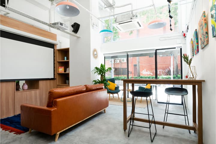 Spacious, high-ceiling common area lounge featuring a brown leather sofa, comfortable accent chairs, a high table with stools, and a large retractable projector screen against the far wall.