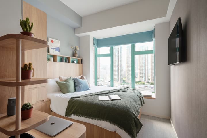 A clean, modern studio apartment bedroom with light wood furniture, a bed with a green throw, a small desk, and a large window overlooking a city skyline.