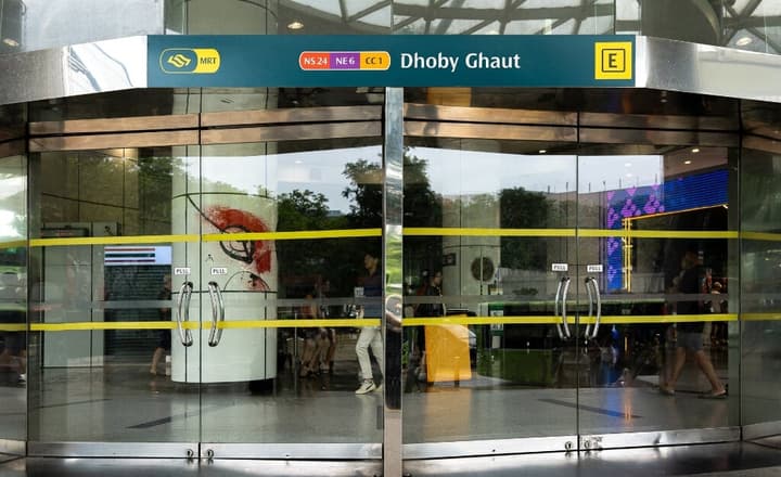 Dhoby Ghaut MRT station entrance through glass doors, with the overhead signage displaying the station name and NS24, NE6, CC1 train lines.