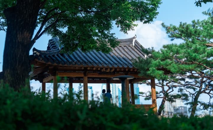 Traditional Korean wooden pavilion (Jeongja) in Seonyudo Park, viewed through lush green foliage, with modern city skyscrapers visible in the background.