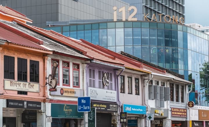 A row of colorful, traditional shophouses with small businesses, contrasting sharply with the modern, glass-facade i12 Katong mall towering behind them.