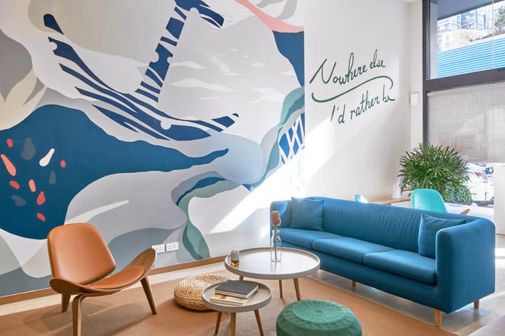 A bright, modern communal lounge featuring a large abstract wall mural with an anchor motif, a blue sofa, a brown leather chair, and the quote, "Nowhere else I'd rather be."