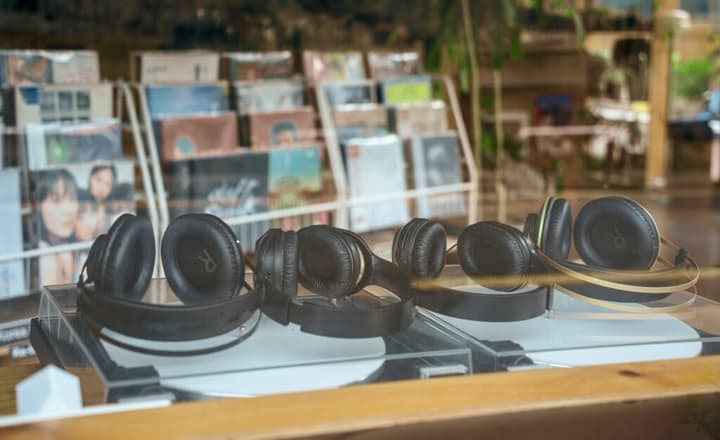 Close-up of several black wireless headphones displayed on clear stands in a window, with a blurred background of stacked vinyl records and album art in a music shop.