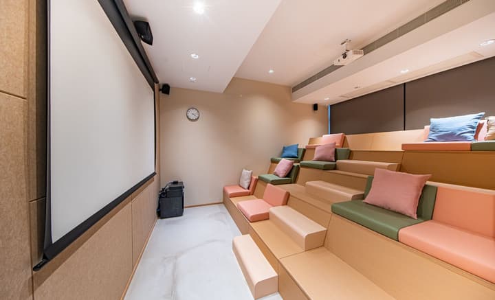 A small screening or movie room with built-in, tiered bench seating with cushions in shades of pink, green, and blue. A large white projection screen is mounted on the left wall, with a ceiling-mounted projector on the opposite side.