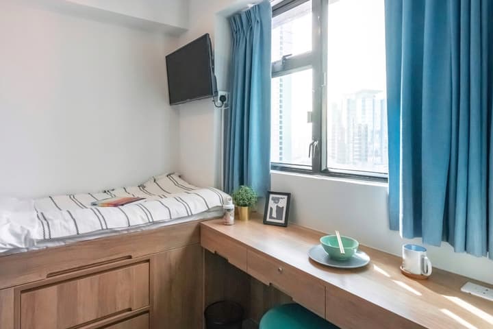 Bright, compact studio bedroom featuring a single bed built into a wooden storage platform, a wooden desk beneath the window, a wall-mounted TV, and bright blue curtains.