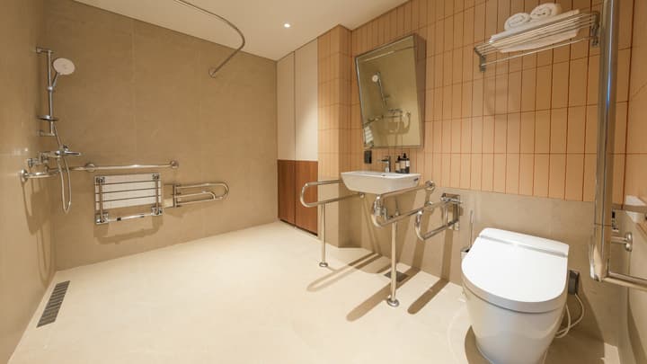 Spacious, accessible apartment bathroom featuring a smart toilet, wall-mounted sink with open space beneath, and a large shower area, all equipped with extensive grab bars and a sleek, modern aesthetic.