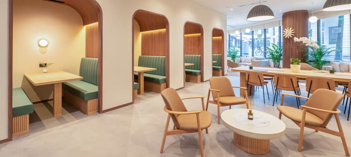 Bright, modern communal lounge and co-working space featuring private arched wooden dining/work booths with green upholstered seating, a long communal table, and a contemporary low seating area with tan chairs.