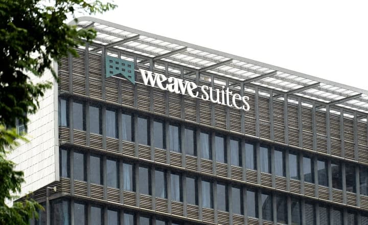 Close-up of the exterior of the Weave Suites building showing the logo and signage against the modern facade with glass windows and horizontal slat paneling.