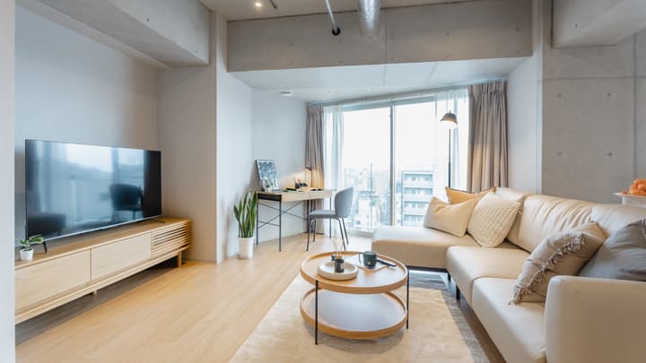 Wide view of a bright apartment living area featuring an L-shaped light tan sofa, a wooden coffee table, a low-profile wood media console, and a large window, with a desk nook visible in the background.
