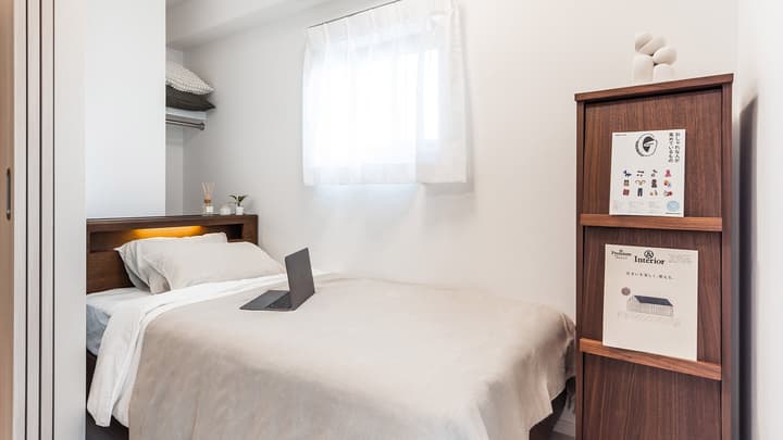A bright, minimalist single bedroom with a wooden bed frame, back-lit headboard, laptop, and a tall, narrow wooden shelving unit.