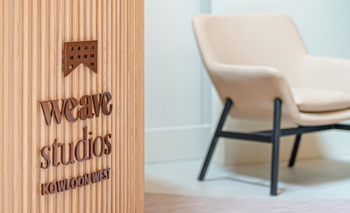 A close-up shot of dimensional bronze-colored text and a logo mounted on a textured, vertically slatted wooden wall. The text reads "weave studios KOWLOON WEST." In the background, a modern, minimalist beige armchair is visible.